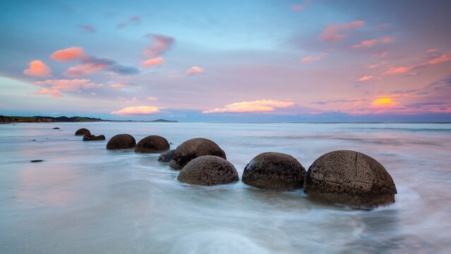 View of the Moeraki Boulders Beach with large spherical boulders resting on the sand at sunset, featuring long exposure waves and a colorful sky in Hampden, Otago Region, New Zealand.