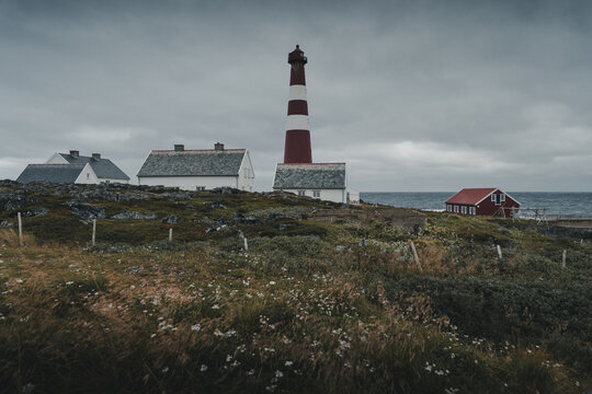 Gamvik, Finnmark, Norway - 26 August 2025: View of Slettnes Lighthouse standing on a rocky coastline with traditional white buildings and a red house under a cloudy sky.