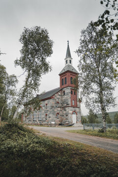 Utsjoki, Finland - 25 August 2025: View of Utsjoki Church, a historic stone building with a red brick tower, surrounded by birch trees and a gravel path under a cloudy sky.