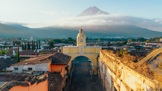 Aerial view of the yellow Santa Catalina Arch clock tower with colonial rooftops and a majestic volcano in the background in Antigua Guatemala, Sacatepequez Department, Guatemala.