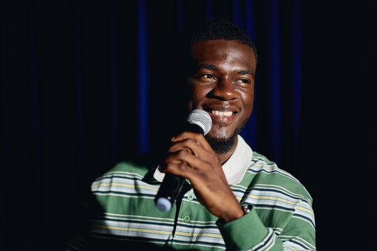 African American young adult male student smiling while holding microphone on theater stage. Image represents public speaking and performing arts education, medium shot