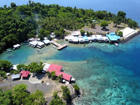 Aerial view of a coastal village with stilt houses over turquoise water, small boats, lush green vegetation, and clear coral reefs along the shoreline in Ternate, North Maluku, Indonesia.