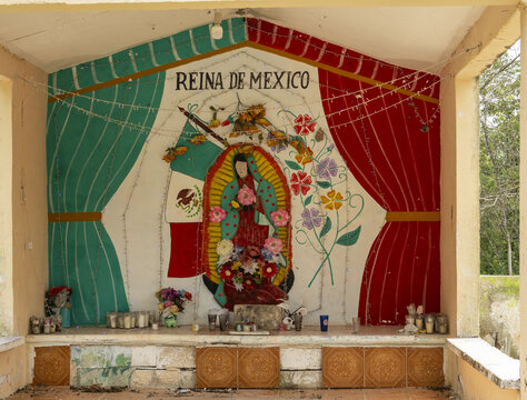 View of a religious shrine featuring a mural of Our Lady of Guadalupe and the Mexican flag, adorned with flowers and candles in Izamal, Yucatan, Mexico.