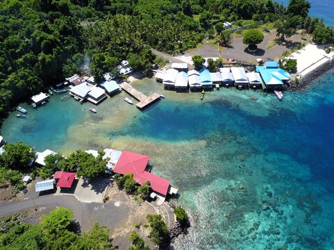 Aerial view of a coastal village with stilt houses over turquoise water, small boats docked at a pier, and lush tropical vegetation along the shoreline in Ternate, North Maluku, Indonesia.