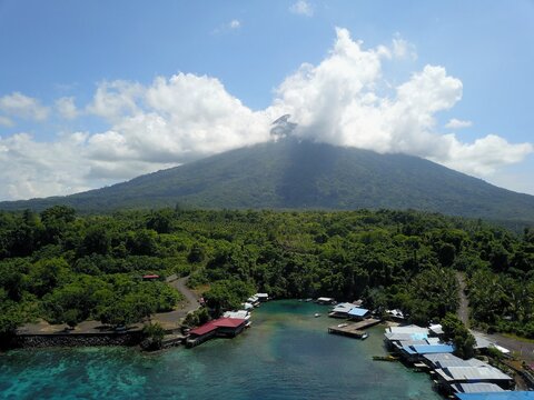 Aerial view of Gamalama Volcano rising above a lush green coastal village with stilt houses and turquoise waters in Ternate, North Maluku, Indonesia.