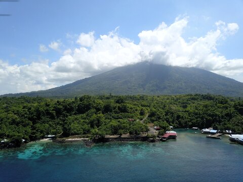 Aerial view of Gamalama volcano rising above lush green forests and the turquoise coastal waters of Ternate, North Maluku, Indonesia.