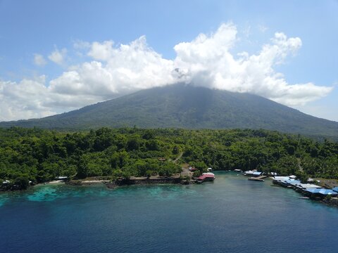 Aerial view of the lush slopes of Gamalama volcano rising above a coastal village with houses on stilts and turquoise waters in Ternate, North Maluku, Indonesia.