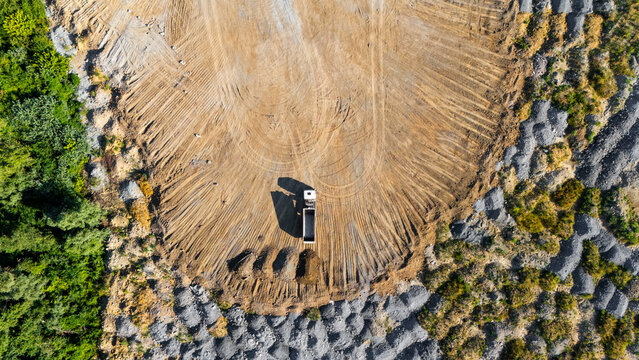 Aerial view of a dump truck parked in a large quarry with piles of gravel, tire tracks on the dirt ground, and surrounding forest in Beocin, Vojvodina, Serbia.