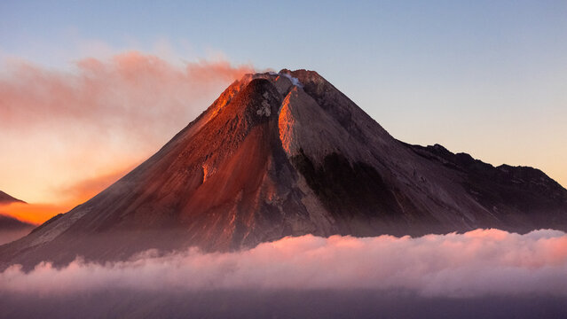 Aerial view of Mount Merapi with volcanic smoke rising above a sea of clouds during golden hour in Kecamatan Pakem, Daerah Istimewa Yogya, Indonesia.