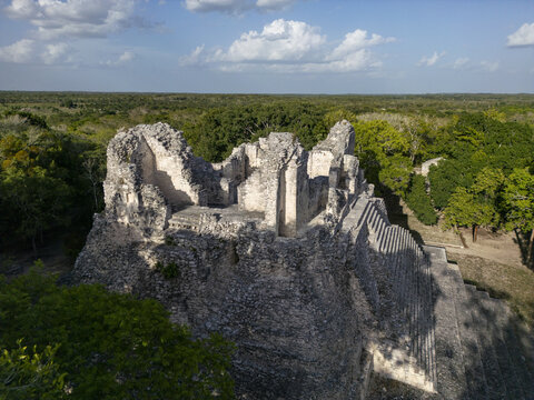 Aerial view of the ancient Mayan ruins of Becan, featuring weathered stone structures and stepped pyramids surrounded by dense jungle canopy under a bright blue sky in Calakmul, Campeche, Mexico.