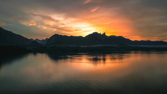 Aerial view of Lake Thun at sunset with the Bernese Alps silhouetted against a vibrant orange sky, reflecting on the calm water in Thun, Canton of Bern, Switzerland.
