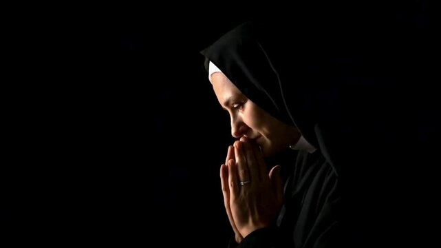 A nun in a black habit prays with hands clasped, eyes closed, and head bowed in a serene, dark setting.	pray video
