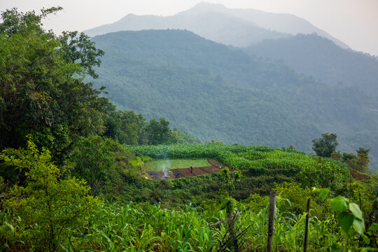 View of a small local village with terraced fields, lush green vegetation, and people working in a field with a small fire against a backdrop of mountains in Pokhara, Gandaki Province, Nepal.