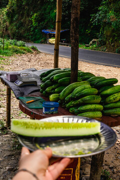 View of a hand holding a sliced cucumber over a wooden table piled with fresh cucumbers at a roadside market stall in a small local village near Pokhara, Gandaki Province, Nepal.