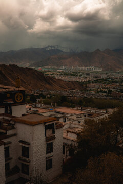 View of the historic white architecture of the Potala Palace overlooking the urban landscape and distant snow-capped mountains under a dramatic cloudy sky in Lhasa, Tibet, China.