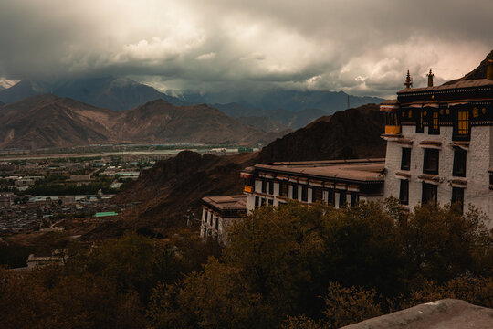 View of the historic Potala Palace with its traditional white and red walls set against a dramatic cloudy sky and the mountainous landscape of Lhasa, Tibet, China.