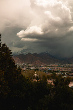 View of the Lhasa cityscape nestled against snow-capped mountains under a dramatic stormy sky with dark clouds and foreground trees in Lhasa, Tibet, China.