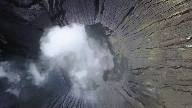 Aerial view of the volcanic crater of Mount Bromo with swirling white smoke rising from the deep, rugged, and textured dark grey walls, Tosari, East Java, Indonesia.