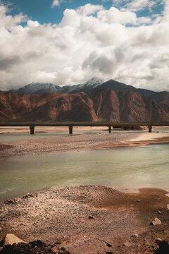 View of a river flowing under a long bridge with snow-capped mountains in the background under a cloudy sky in Tibet, China.