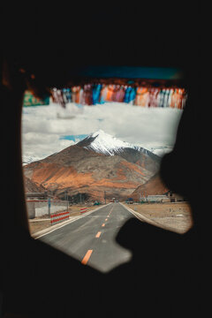 View of a paved road stretching towards a snow-capped mountain peak under a cloudy sky, seen through a vehicle window in Tibet, China.