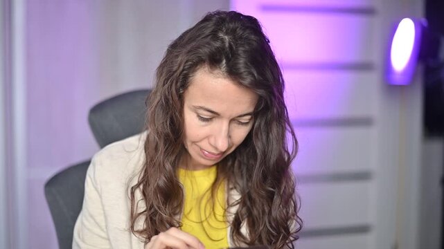 Focused curly haired woman using her smartphone typing and scrolling social media feed in her modern home office at night