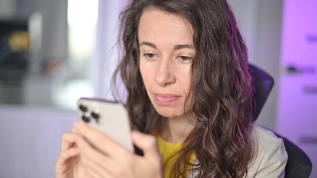 Focused curly haired woman using her smartphone typing and scrolling social media feed in her modern office at night home