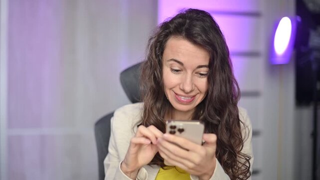 Focused curly haired woman using her smartphone typing and scrolling social media feed in her modern at night home office