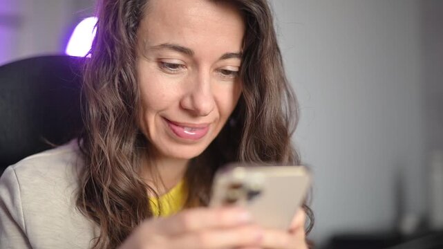 Focused curly haired woman using her smartphone typing and scrolling social media feed in her modern night home office