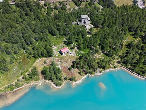 Aerial view of Wasserkraftwerk Palu hydroelectric power plant near the turquoise Lago di Palu, surrounded by dense pine forests and rugged alpine terrain in Poschiavo, Grisons, Switzerland.