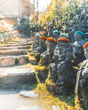 View of stone Jizo statues wearing colorful knitted caps lined along a mossy stone staircase in a temple garden in Japan.