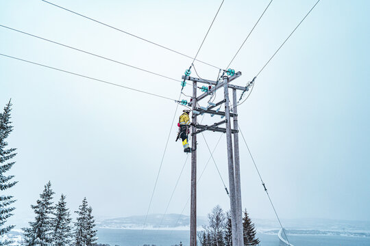 Tromso, Troms, Norway - 05 February 2020: View of a worker in high-visibility gear climbing a wooden power line pole in a snowy winter landscape.