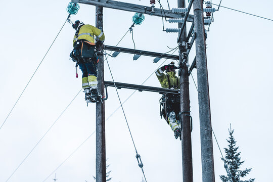 Tromso, Troms, Norway - 05 February 2020: View of two electrical workers in high-visibility gear climbing wooden utility poles to perform maintenance on power lines in winter.