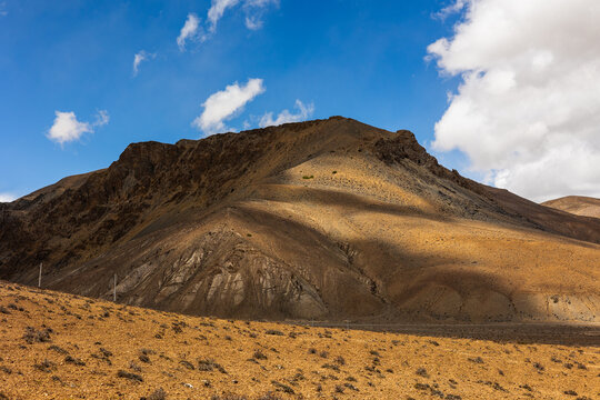View of the rugged arid mountains near Peiku Cuo in the high-altitude landscape under a bright blue sky with scattered clouds in Shigatse, Tibet, China.