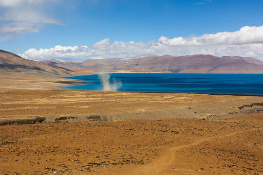 View of Peiku Cuo alpine lake with a dust devil rising from the arid shoreline against a backdrop of mountains in Shigatse, Tibet, China.