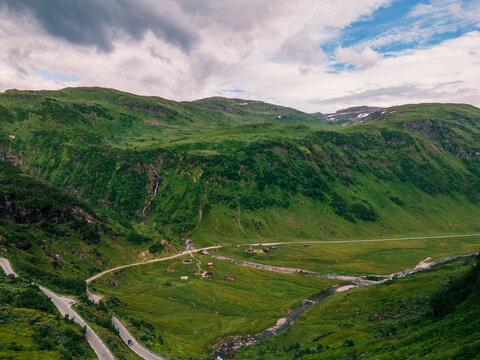 Aerial view of a winding road through a lush green mountain valley with a small stream and scattered cabins in Norway.