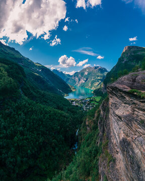 Aerial view of Geirangerfjord surrounded by steep mountains, lush green forests, and deep blue water under a bright sky in Stranda Municipality, More og Romsdal, Norway.
