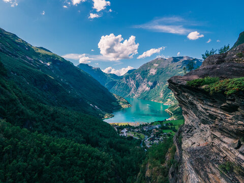 Aerial view of Geirangerfjord surrounded by steep mountains, lush green forests, and deep blue water under a bright sky in Stranda Municipality, More og Romsdal, Norway.