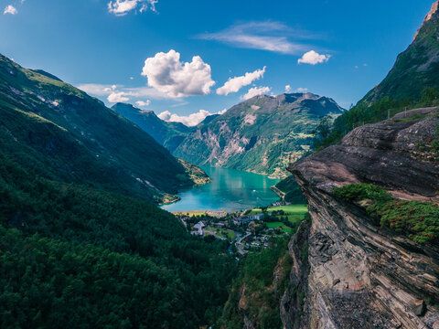 Aerial view of Geirangerfjord surrounded by steep mountains, lush forests, and a small village nestled in the valley under a blue sky in Stranda Municipality, More og Romsdal, Norway.