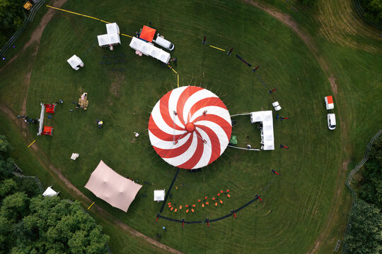 Aerial view of a large red and white striped circus tent set up in a lush green park surrounded by smaller white tents and trees in Vilnius, Vilnius County, Lithuania.