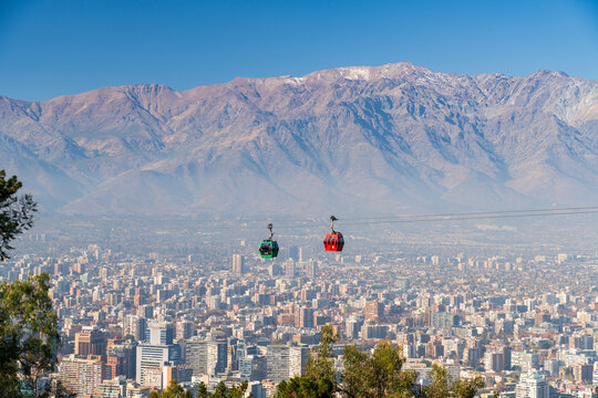 View of the red and green cabins of the Cerro San Cristobal Cable Car moving above the urban skyline of Santiago with the Andes mountains in the background in Providencia, Region Metropolitana, Chile.