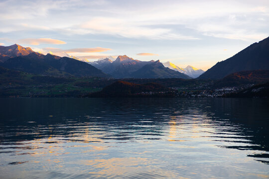 View of the sunset over Lake Thun with golden light reflecting on the calm water and the surrounding mountain peaks in Thun, Canton of Bern, Switzerland.