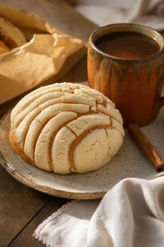 Mexican concha sweet bread on a plate with a clay mug of coffee. Traditional pan dulce breakfast with a cinnamon stick on a wooden table