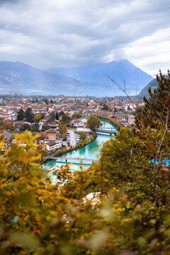 View of Harder Kulm Mountain overlooking the town of Unterseen with the Aare River, surrounded by autumn trees and mountains under a cloudy sky in Unterseen, Canton of Bern, Switzerland.