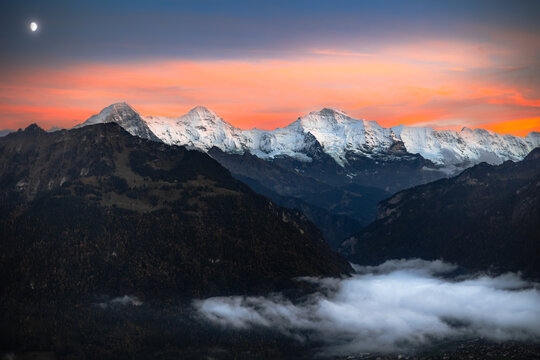 View of the snow-capped Eiger, Monch, and Jungfrau peaks at sunset from Harder Kulm with low-hanging clouds and a crescent moon in the sky, Unterseen, Canton of Bern, Switzerland.