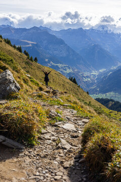 View of a hiker standing with arms raised on a rocky mountain trail overlooking the Niesen mountain range and the valley in Reichenbach im Kandertal, Canton of Bern, Switzerland.