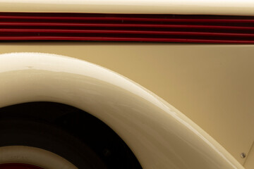 Close-up view of a cream colored classic car showing the curved wheel arch and part of the tire, with a horizontal red painted grille strip above. Smooth © Viktar
