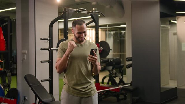 Excited African American man athlete celebrating online phone victory in gym happy male holding smartphone winning prize fitness trainer surprised good news achievement sports betting success reaction