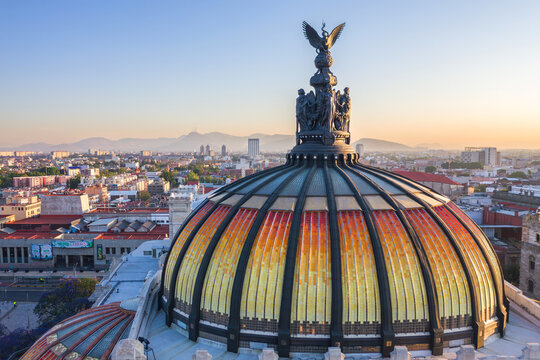 Aerial view of the iconic orange and yellow tiled dome of the Palacio de Bellas Artes, topped with a bronze sculpture, overlooking the urban cityscape of Ciudad de Mexico, Mexico.