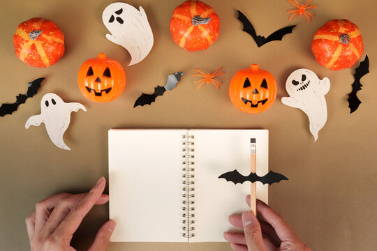 Cropped hands with pencil and open blank notebook on brown background with Halloween decorations