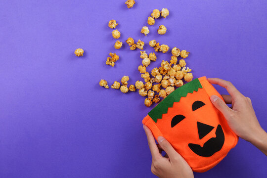 Cropped hands holding jack o lantern pumpkin bag with caramel popcorn on purple background for Halloween